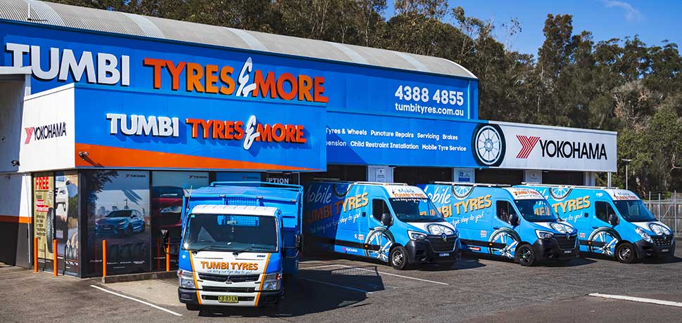 Exterior view of Tumbi Tyres & More store with branded vehicles parked in front.